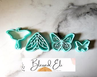 May include: Four turquoise blue plastic cookie cutters shaped like butterflies and moths. The cutters are arranged in a row on a white marble surface. The text "Blessed Eli Polymer Clay Jewelry" is visible in the bottom of the image.