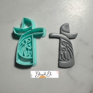 May include: Two cross-shaped cookie cutters. One is a light teal color, and the other is gray. Both have a detailed design of a religious scene. The text "Blessed Eli" is at the bottom.