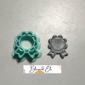 May include: Two award-shaped cookie cutters. One is a light teal colour, and the other is a grey colour. The teal cutter is a 3D printed plastic. The grey cutter is a flat, solid shape. The text "Blessed Eli" is printed on a white and pink background.