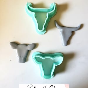 Könnte beinhalten: Satz mit vier Plastik-Ausstechern in Form von Kuh- und Bullenköpfen. Zwei Ausstecher sind hellblau und zwei sind grau. Die Ausstecher sind auf einer weißen Oberfläche angeordnet. Der Text "Blessed Eli Polymer Clay Jewelry" ist in der rechten unteren Ecke des Bildes sichtbar.