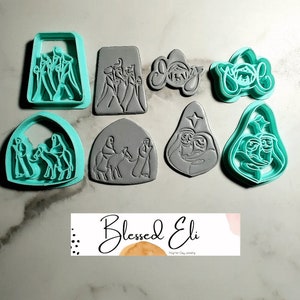 Puede incluir: Un conjunto de ocho cortadores de galletas de plástico gris y azul verdoso que representan diferentes escenas navideñas, incluyendo los tres reyes magos, un pesebre y las palabras "Alegría" y "Navidad".