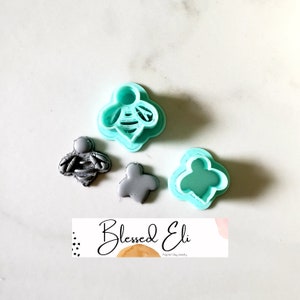 May include: A set of three polymer clay cutters, two are bee-shaped and one is a flower shape. The cutters are light blue and gray. The cutters are on a white surface with a business card that says "Blessed Eli Polymer Clay Jewelry".