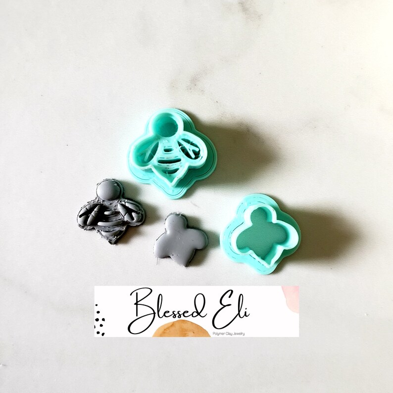 May include: A set of three polymer clay cutters, two are bee-shaped and one is a flower shape. The cutters are light blue and gray. The cutters are on a white surface with a business card that says "Blessed Eli Polymer Clay Jewelry".