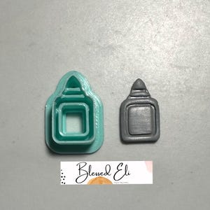 May include: A teal polymer clay cutter and a grey polymer clay charm shaped like a bottle. The cutter has a rectangular form with a rounded top and a square cutout. The charm has a similar shape with a textured surface. The image includes the text "Blessed Eli".