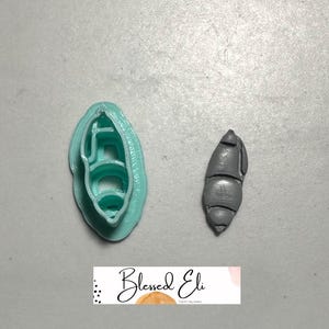 May include: A light teal polymer clay cutter and a grey polymer clay creation. The cutter is in the shape of a shell. The grey creation is a shell. The text "Blessed Eli" is at the bottom.