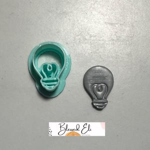 May include: A light blue, plastic lightbulb-shaped cookie cutter next to a grey lightbulb-shaped object. The cookie cutter has a detailed design. The grey object has a textured surface. The image includes the text "Blessed Eli".