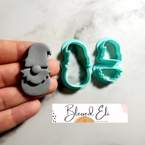 Puede incluir: Un cortador de galletas de plástico gris en forma de gnomo con barba y sombrero. El cortador se muestra junto a un cortador de galletas de plástico azul turquesa en dos piezas, una para el cuerpo y otra para la barba.