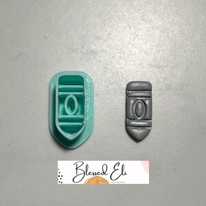 May include: A teal polymer clay cutter and a grey polymer clay pendant. The cutter is shield-shaped with an oval in the centre. The pendant is a miniature version of the cutter. The image includes the text "Blessed Eli".