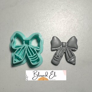 May include: A teal bow-shaped cookie cutter and a grey polymer clay bow. The cutter is plastic and detailed. The clay bow has a textured surface and realistic folds. The image includes the text "Blessed Eli". The measurements are in centimeters.