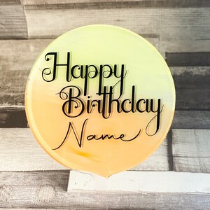 Può includere: Un topper per torta rotondo, giallo e arancione con il testo "Happy Birthday Name" in lettere corsive nere.