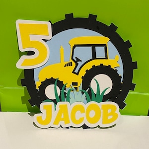 May include: Yellow tractor cake topper with a black gear wheel, green grass, and the number 5 and the name Jacob in yellow letters.