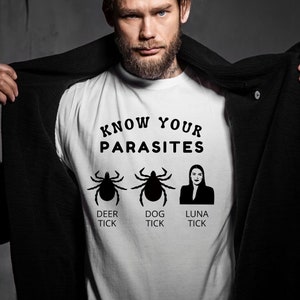 Puede incluir: Camiseta blanca con texto negro que dice "Know Your Parasites". Tres siluetas negras están debajo del texto: una garrapata de venado, una garrapata de perro y una mujer con el texto "Luna Tick" debajo.