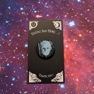 Könnte beinhalten: Ein schwarz-weißer Knopf mit einem blauen und weißen Bild eines Gesichts mit einem Gittermuster. Der Knopf befindet sich auf einer schwarzen Karte mit dem Text "Needle Say More...?" und "Thank You!" in weiß.