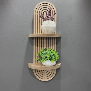 May include: A wooden wall shelf with a rounded arch design. The shelf has two tiers and features a light brown wood finish. The top tier has a small white pot with purple flowers, and the bottom tier has a small white pot with green plants.