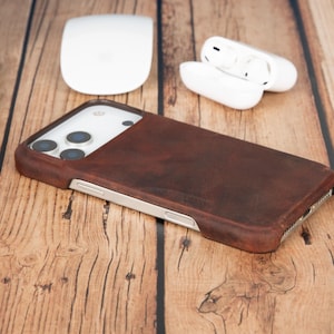 Puede incluir: Una funda de cuero marrón oscuro para smartphone con una textura suave y mate que cubre la parte trasera y los laterales, con recortes precisos para las lentes de la cámara y los botones laterales, diseñada para un smartphone moderno.
