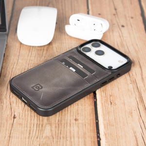 Può includere: Una custodia per smartphone grigio scuro con uno scomparto per carte integrato sul retro, dotata di una cornice nera e un logo visibile. La custodia presenta una texture liscia simile alla pelle e include ritagli per lenti e pulsanti.