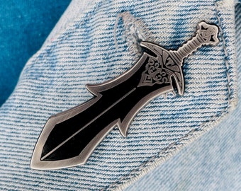 May include: A silver and black sword pin with intricate details. The pin is attached to a denim jacket.