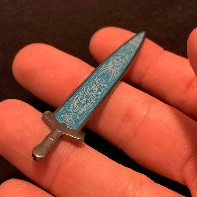 May include: A silver-toned sword pin with a blue enamel design. The sword has a detailed design with intricate patterns and a pointed tip.