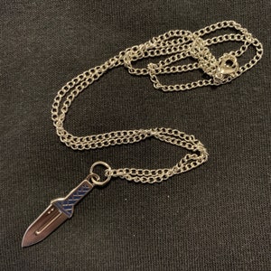 Puede incluir: Un collar de cadena de plata con un colgante de cuchillo de plata. El cuchillo tiene una hoja texturizada y un mango de plata.