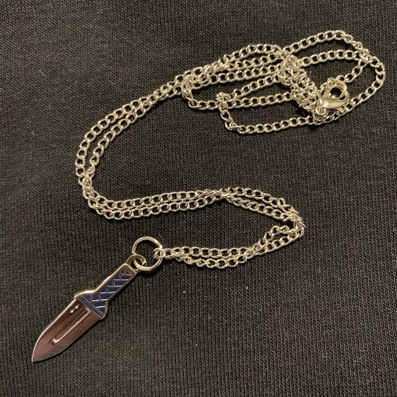 Puede incluir: Un collar de cadena de plata con un colgante de cuchillo de plata. El cuchillo tiene una hoja texturizada y un mango de plata.