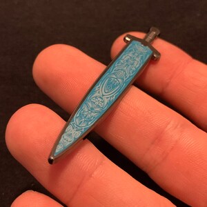 May include: A silver-toned sword pin with a blue enamel design. The sword has a detailed design with intricate patterns and a pointed tip.