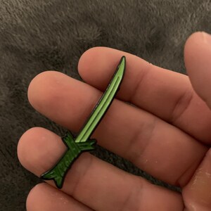 May include: A green enamel pin shaped like a sword. The sword has a black outline and a silver handle.