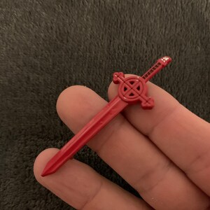 May include: A red plastic sword with a circular design on the handle. The sword is about 5 inches long.