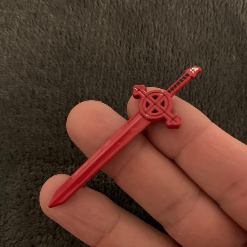 May include: A red plastic sword with a circular design on the handle. The sword is about 5 inches long.