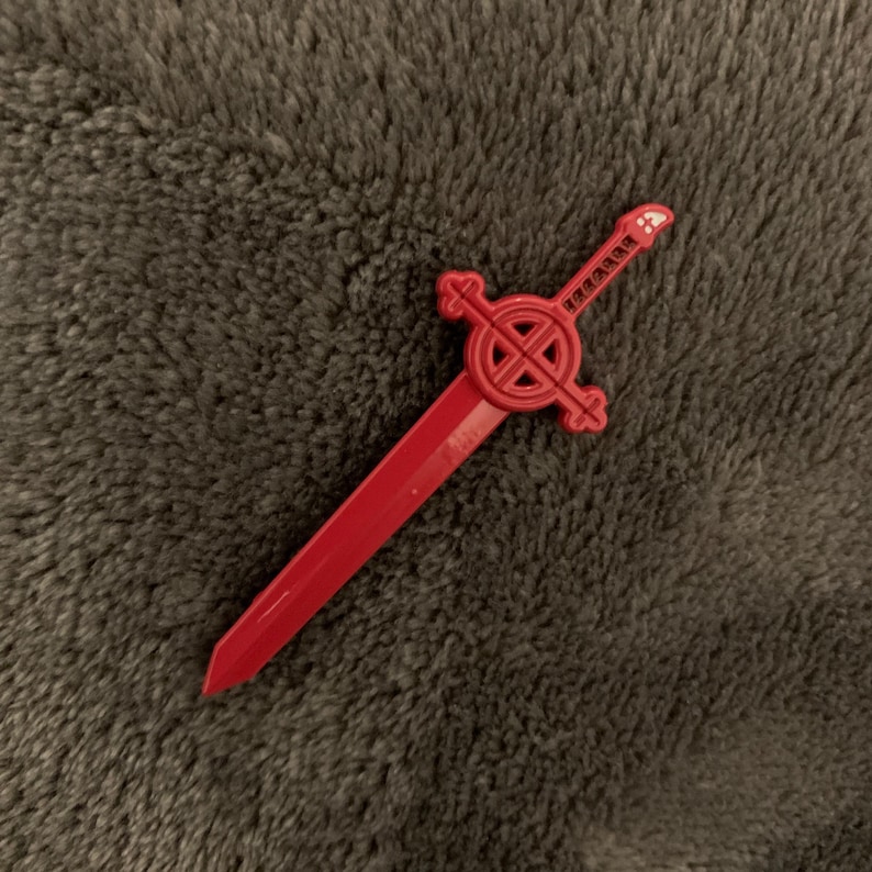 May include: A red plastic sword with a circular design and a cross at the top. The sword is about 10 inches long.