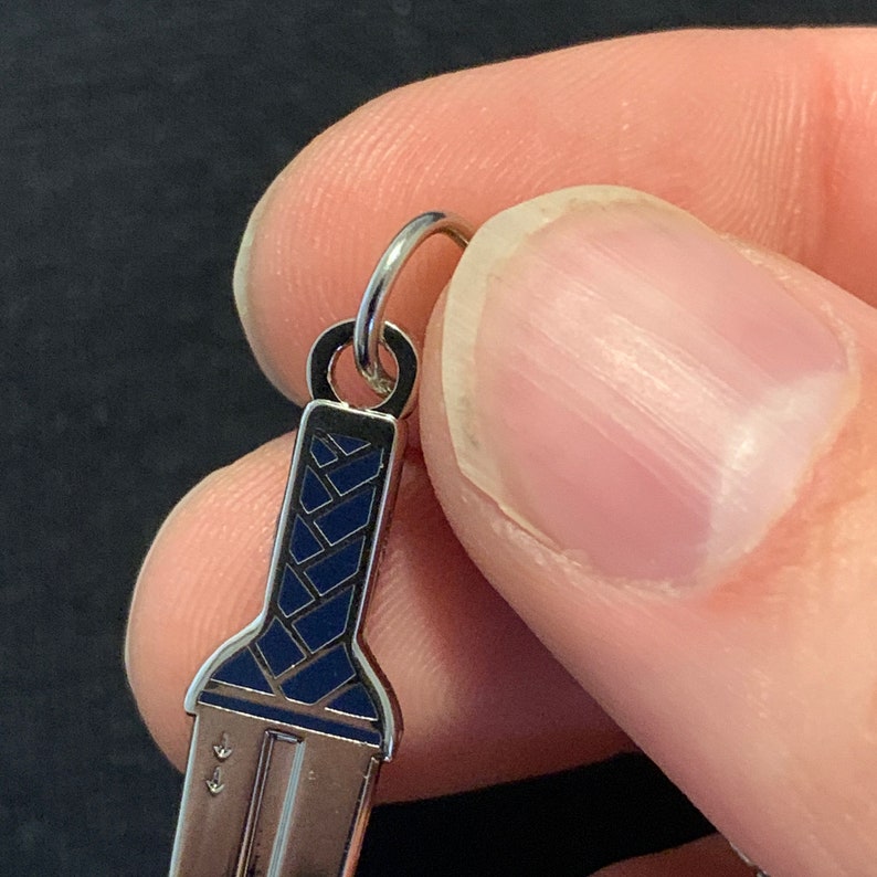Puede incluir: Un amuleto de plata con forma de espada con un mango de esmalte azul. El amuleto tiene un bucle en la parte superior para sujetarlo a una pulsera o collar.