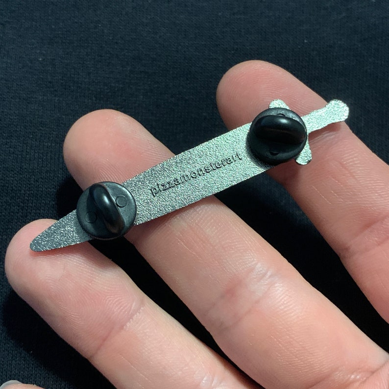 May include: A silver sword pin with a black enamel finish. The back of the pin has the text "pizzamonsterart" printed on it.