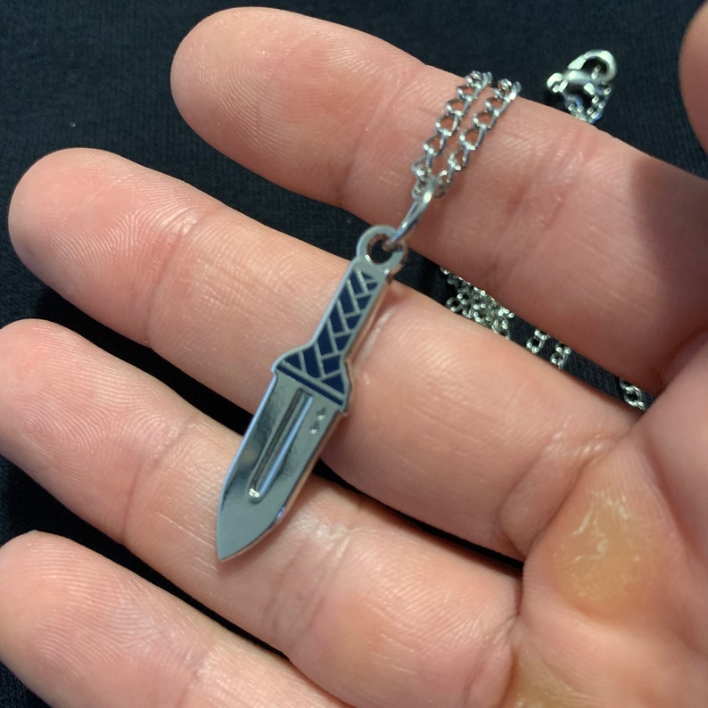 Puede incluir: Un collar de cadena de plata con un colgante de daga de plata. La daga tiene un dise&ntilde;o de esmalte azul en el mango.