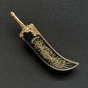 May include: A black and gold sword pin with a detailed design. The sword has a curved blade and a decorative handle.