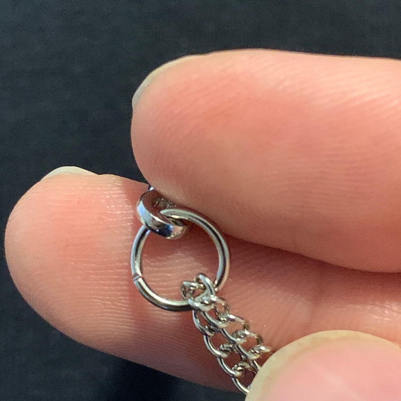 Puede incluir: Primer plano de una cadena de plata con un anillo de salto de plata. El anillo de salto est&aacute; abierto y listo para ser unido a otra pieza de joyer&iacute;a.