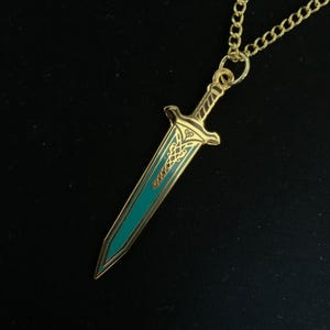 Puede incluir: Un collar con un colgante de espada de color dorado, la hoja es de color turquesa y tiene detalles intrincados. El colgante está unido a una cadena dorada.