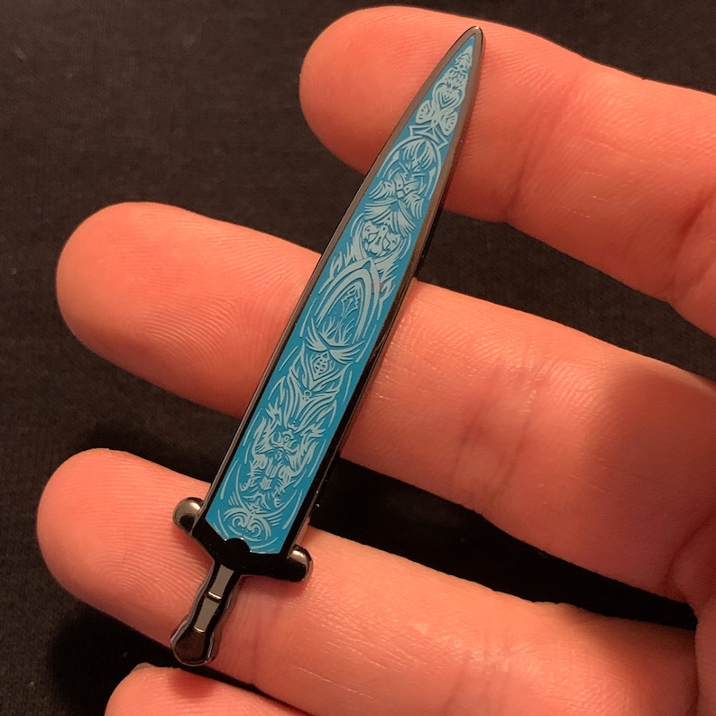 May include: A black metal sword pin with a blue enamel design. The sword has a detailed design with intricate patterns and a pointed tip.