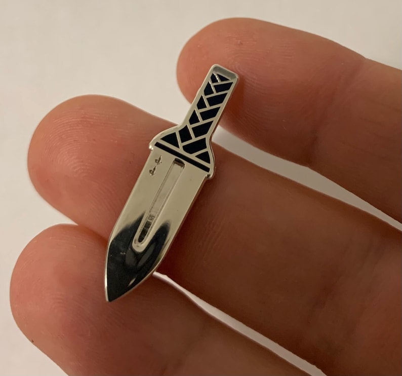 Pu&ograve; includere: Spilla a forma di coltello in metallo argentato con un design in smalto nero. Il coltello ha una punta appuntita e un manico nero con un motivo geometrico.
