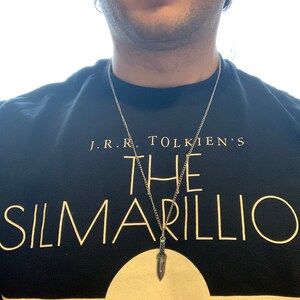 Puede incluir: Un collar de cadena de plata con un colgante de daga de plata. El collar lo lleva una persona que lleva una camiseta negra con el texto "J.R.R. TOLKIEN'S THE SILMARILLION" impreso en ella.