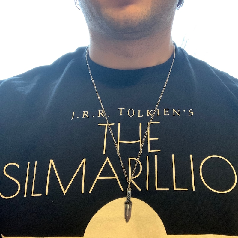 Puede incluir: Un collar de cadena de plata con un colgante de daga de plata. El collar lo lleva una persona que lleva una camiseta negra con el texto "J.R.R. TOLKIEN'S THE SILMARILLION" impreso en ella.