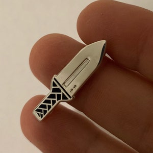 Pu&ograve; includere: Uno spillo color argento a forma di coltello con un manico a motivi bianco e nero.
