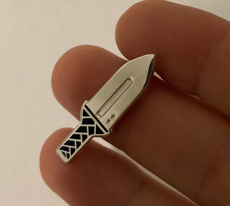 Pu&ograve; includere: Uno spillo color argento a forma di coltello con un manico a motivi bianco e nero.