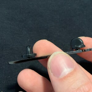 May include: Close-up of the back of a black enamel pin with two metal posts. The posts are used to attach the pin to fabric.