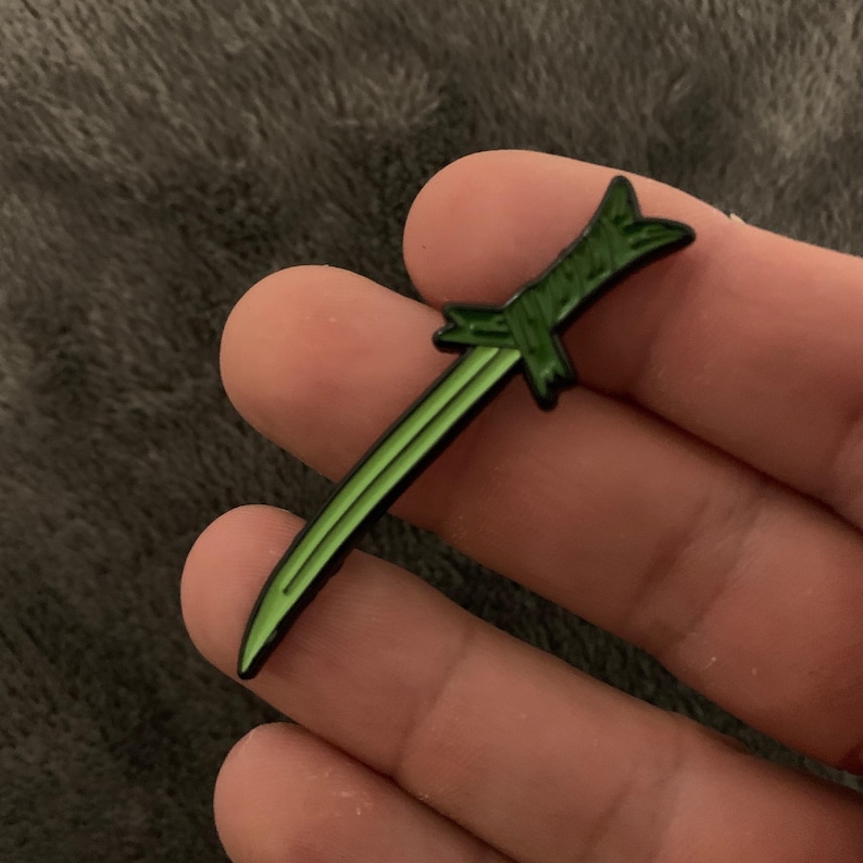 May include: A green and black enamel pin shaped like a sword. The sword is curved and has a sharp point.