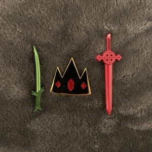 May include: Three enamel pins featuring a black crown with red gems, a green sword, and a red sword with a gold cross.