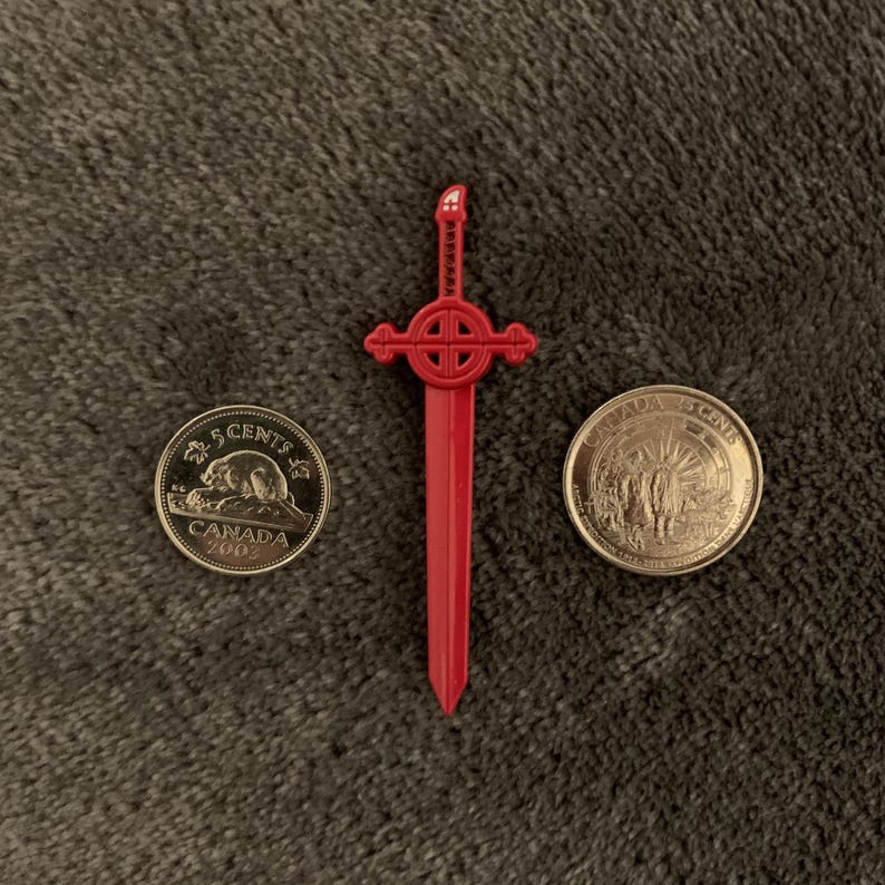 May include: A red plastic sword with a cross-shaped handle and a gold Canadian 5-cent coin from 2002.