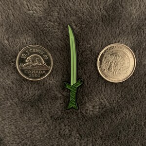May include: A green enamel pin shaped like a katana sword with a black and green handle. The pin is next to two Canadian 5-cent coins, one from 2003 and the other with the text "CANADA CENTS".