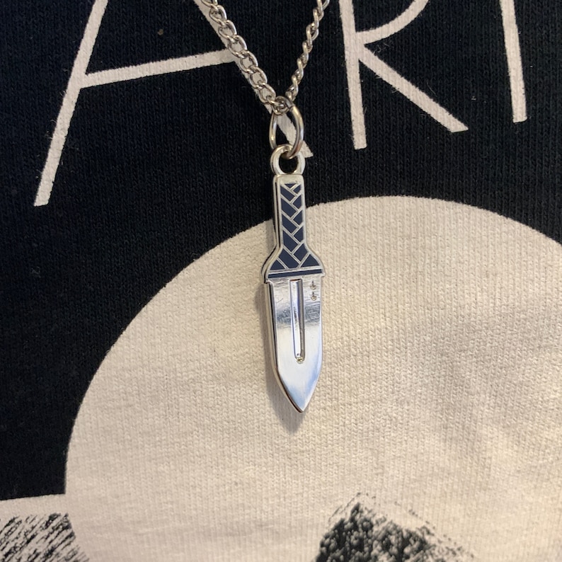 Puede incluir: Un collar de plata con un colgante de daga de plata. La daga tiene un dise&ntilde;o de esmalte negro en el mango.