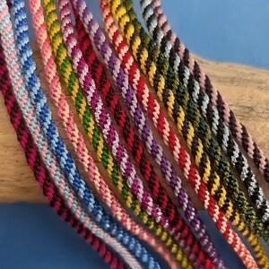May include: A collection of multiple braided cord bracelets featuring various two-tone colour combinations including blue, pink, green, yellow, red, purple, and black. Each bracelet is constructed from twisted thread in a diagonal pattern.