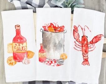Könnte beinhalten: Drei weiße Geschirrtücher mit Aquarell-Illustrationen von Meeresfrüchten. Das erste Handtuch zeigt eine Flasche scharfe Sauce, eine Zitrone, einen Maiskolben und eine Knoblauchzehe. Das zweite Handtuch zeigt einen Topf mit Hummer mit einer Flasche scharfe Sauce und Zitronen. Das dritte Handtuch zeigt einen einzelnen Hummer mit erhobenen Scheren.