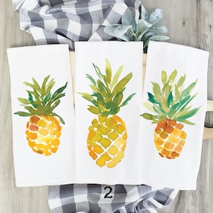 May include: Three white kitchen towels with watercolor pineapple designs. The pineapples are yellow with green leaves and are painted in a loose, abstract style.